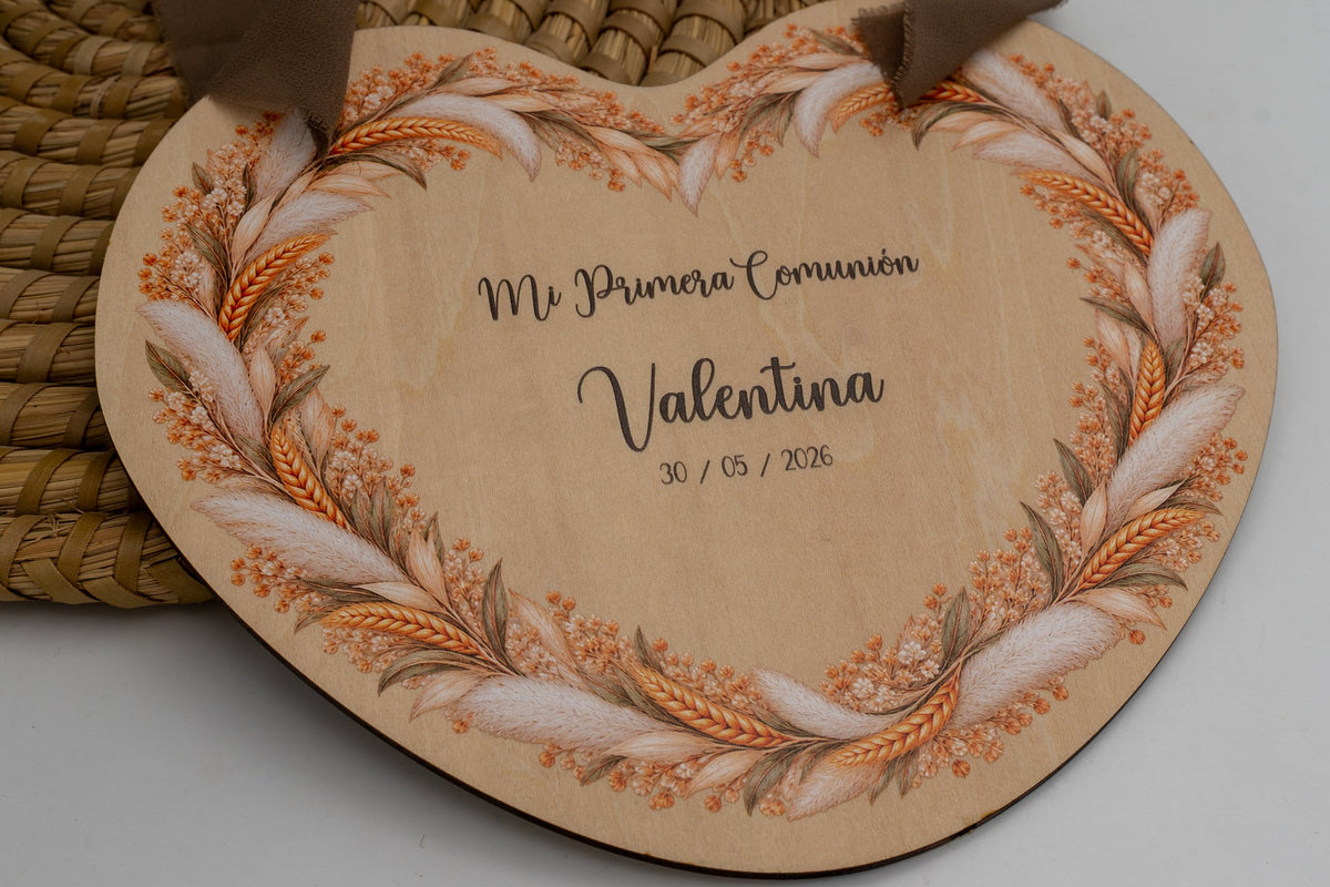 Cartello per la comunione in legno a forma di cuore con corona di spighe ed erba della pampa, con il testo «Mi Primera Comunión», il nome di esempio Valentina e la data, appeso con un nastro marrone.