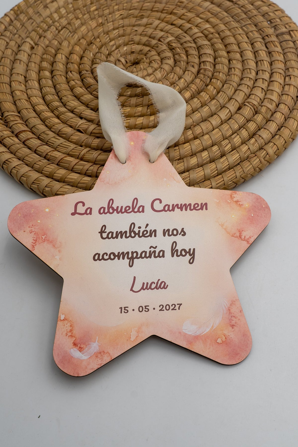 Stella per la comunione con sfondo ad acquerello rosa e piume bianche, la frase «La abuela Carmen también nos acompaña hoy», il nome di esempio Lucía e la data, con nastro color crema.