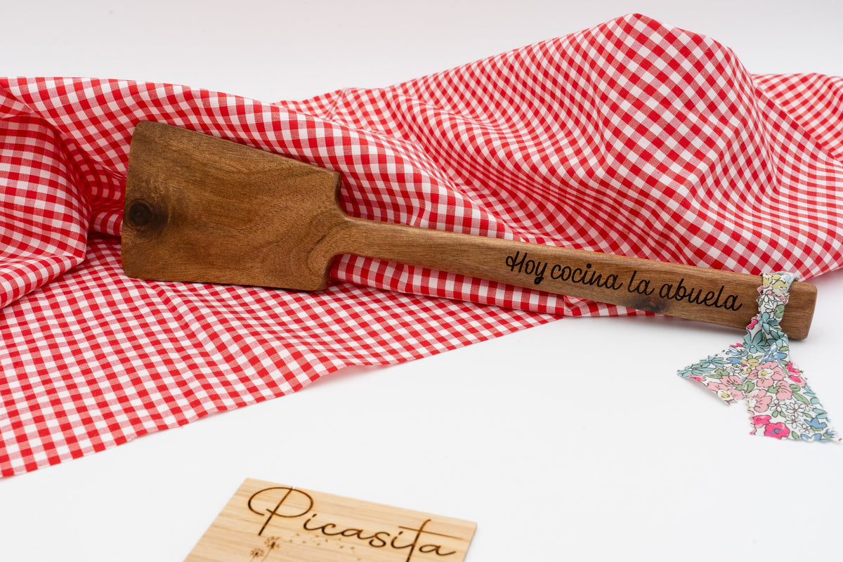 Espátula de madera oscura con mensaje Hoy cocina la abuela sobre mantel de cuadros rojos.
