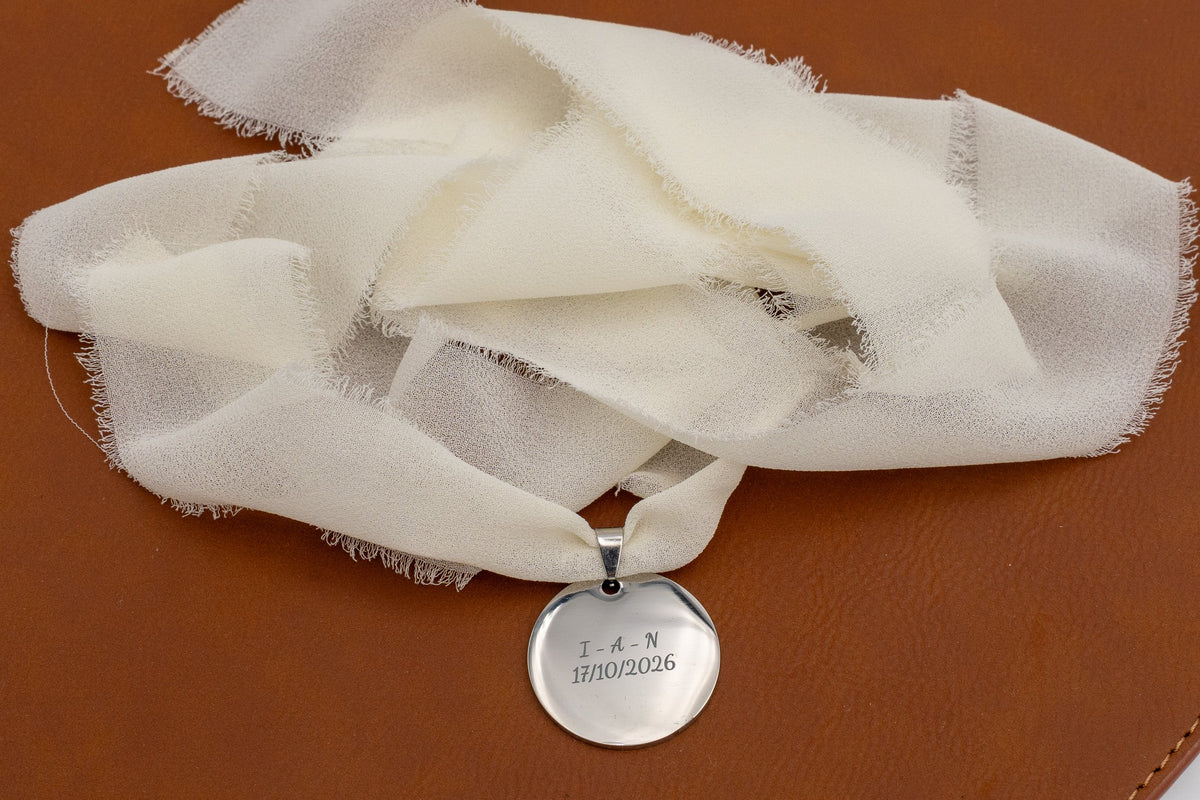 Ciondolo argentato con iniziali e data incise su un nastro di chiffon avorio