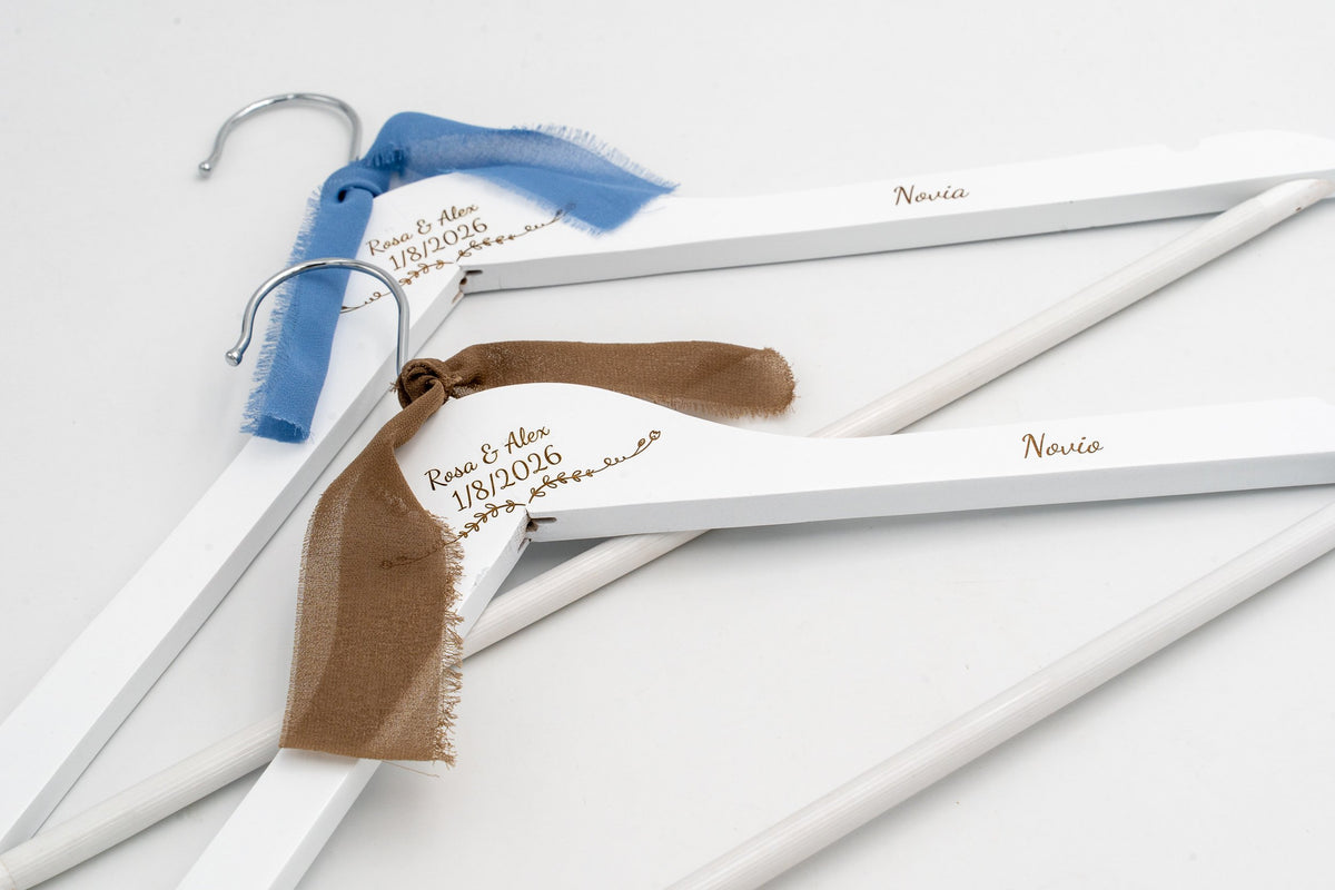 Due grucce bianche per sposi con nomi e data incisi e fiocchi di chiffon azzurri e marroni