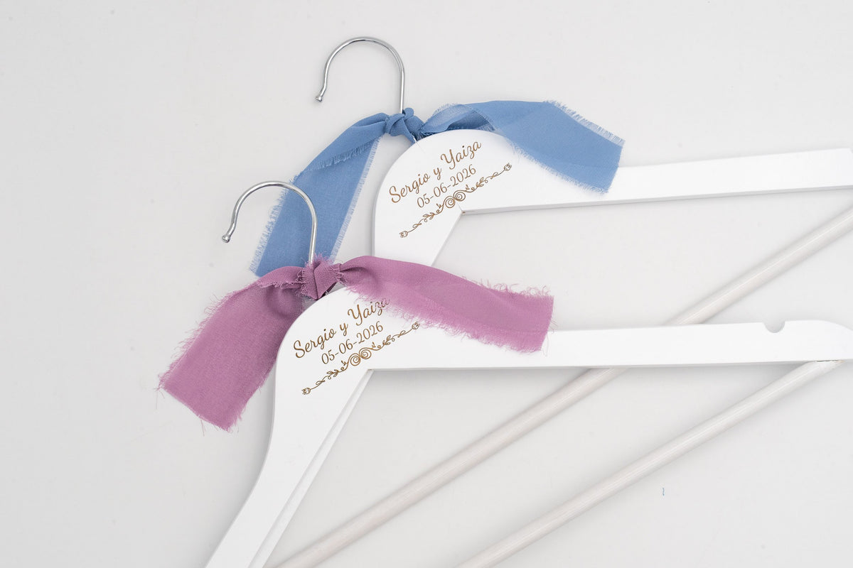Due grucce da matrimonio bianche con i nomi degli sposi incisi e fiocchi di chiffon azzurri e lilla